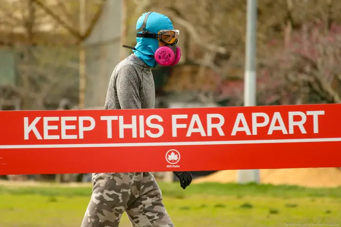 A photo of a person in a mask walking behind a "Keep this far away" sign in a park